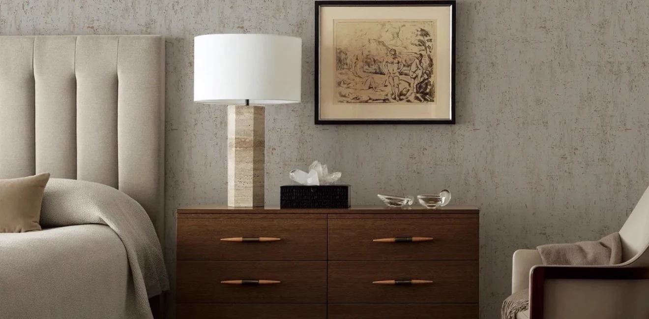 Bedroom with a pale grey cork wallcovering behind a walnut chest of drawers, a travertine table lamp, a framed sketch and an upholstered headboard