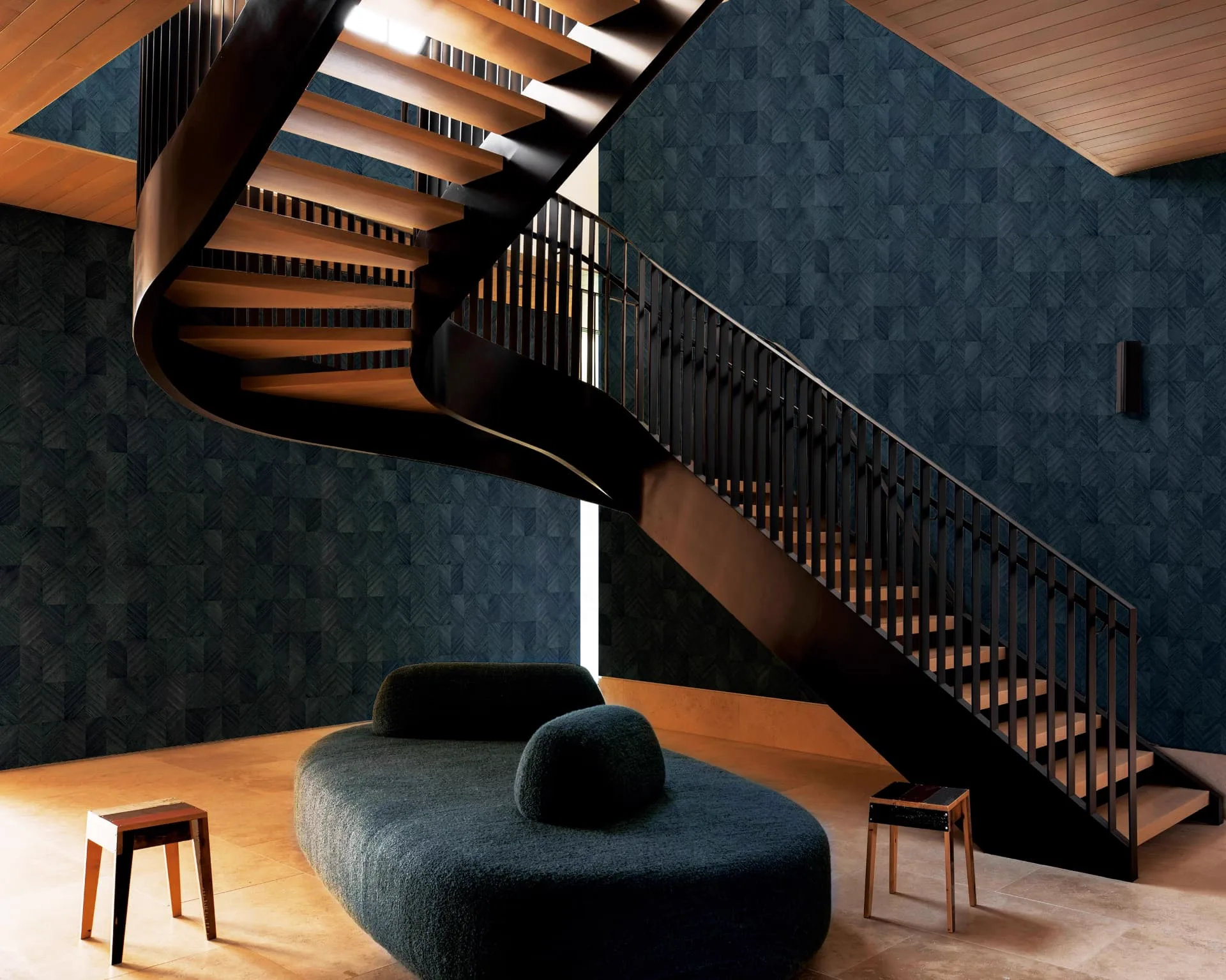 Double-height interior with a deep blue patterned wood veneer wallcovering behind a sculptural dark timber staircase and a curved green upholstered bench