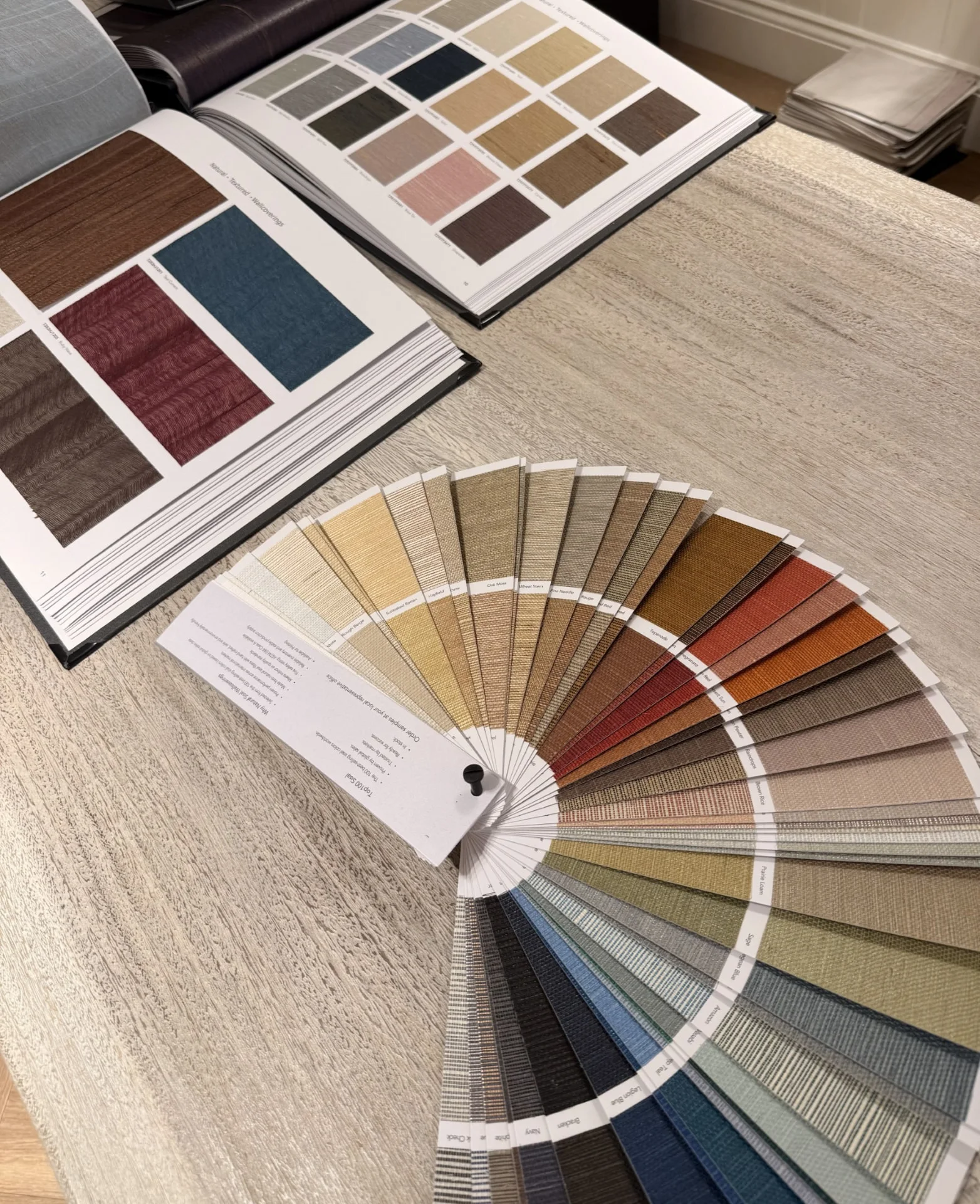Open wallcovering sample books and a fanned colour deck of natural textured swatches laid out on a pale wood table