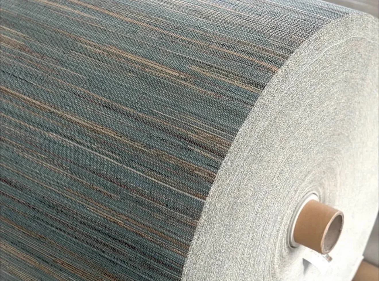 A roll of woven hemp wallcovering in blue-grey and warm tan strands, showing the fibre surface and the rolled edge on its card core