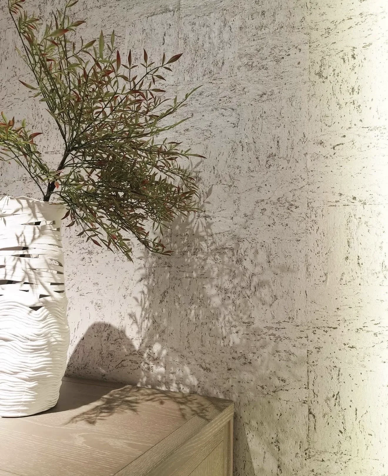 Pale white cork wallcovering with natural cork markings, softly shadowed by a branch in a white woven vase on a light oak cabinet